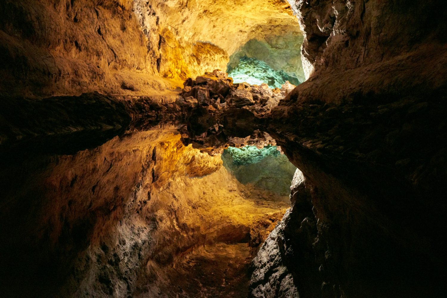 A volcanic lava-tube cave lit gold, its still pool mirroring the rock around a window of turquoise light.